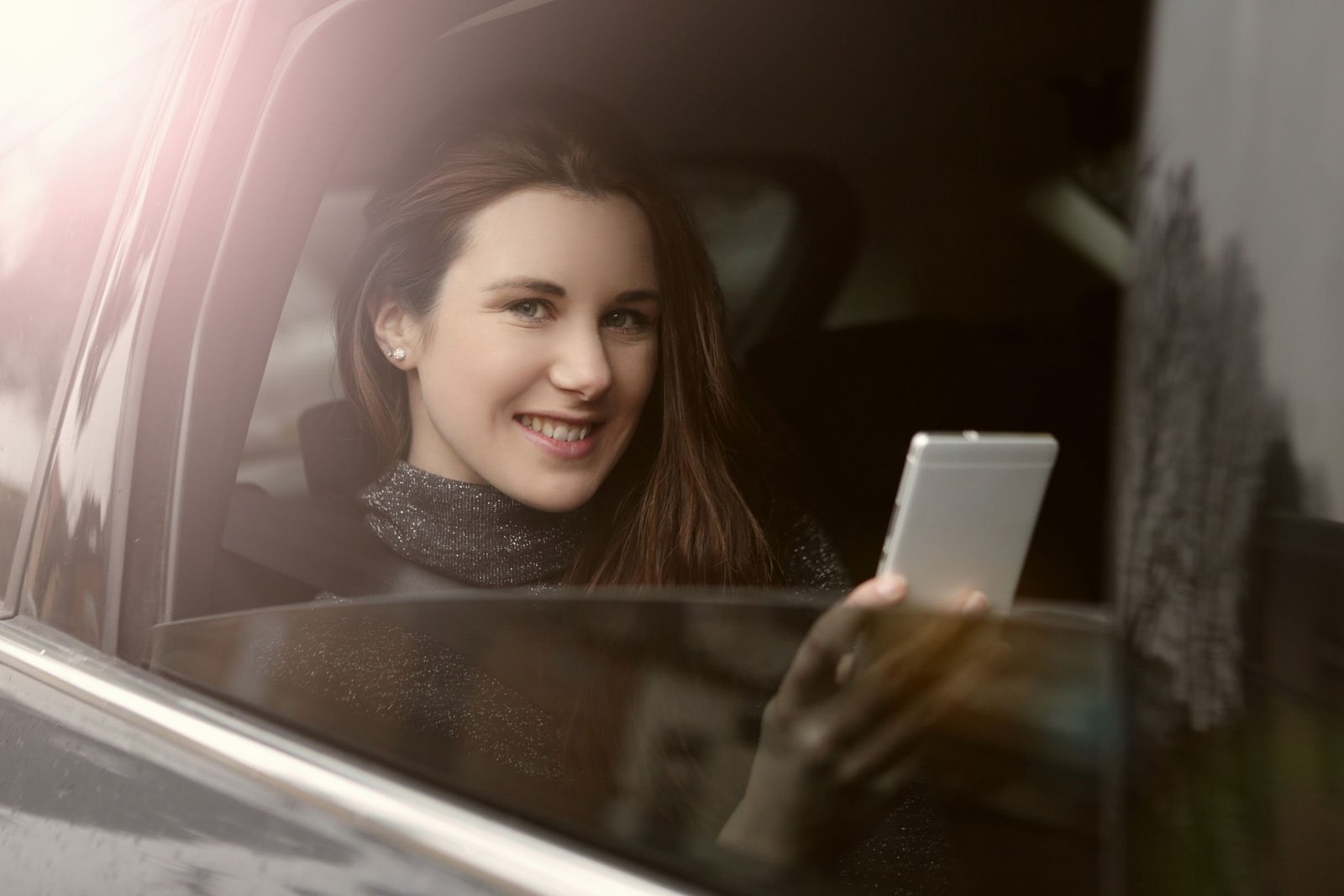 Junge Frau sitzt im Auto und schaut lächelnd vom Handy auf